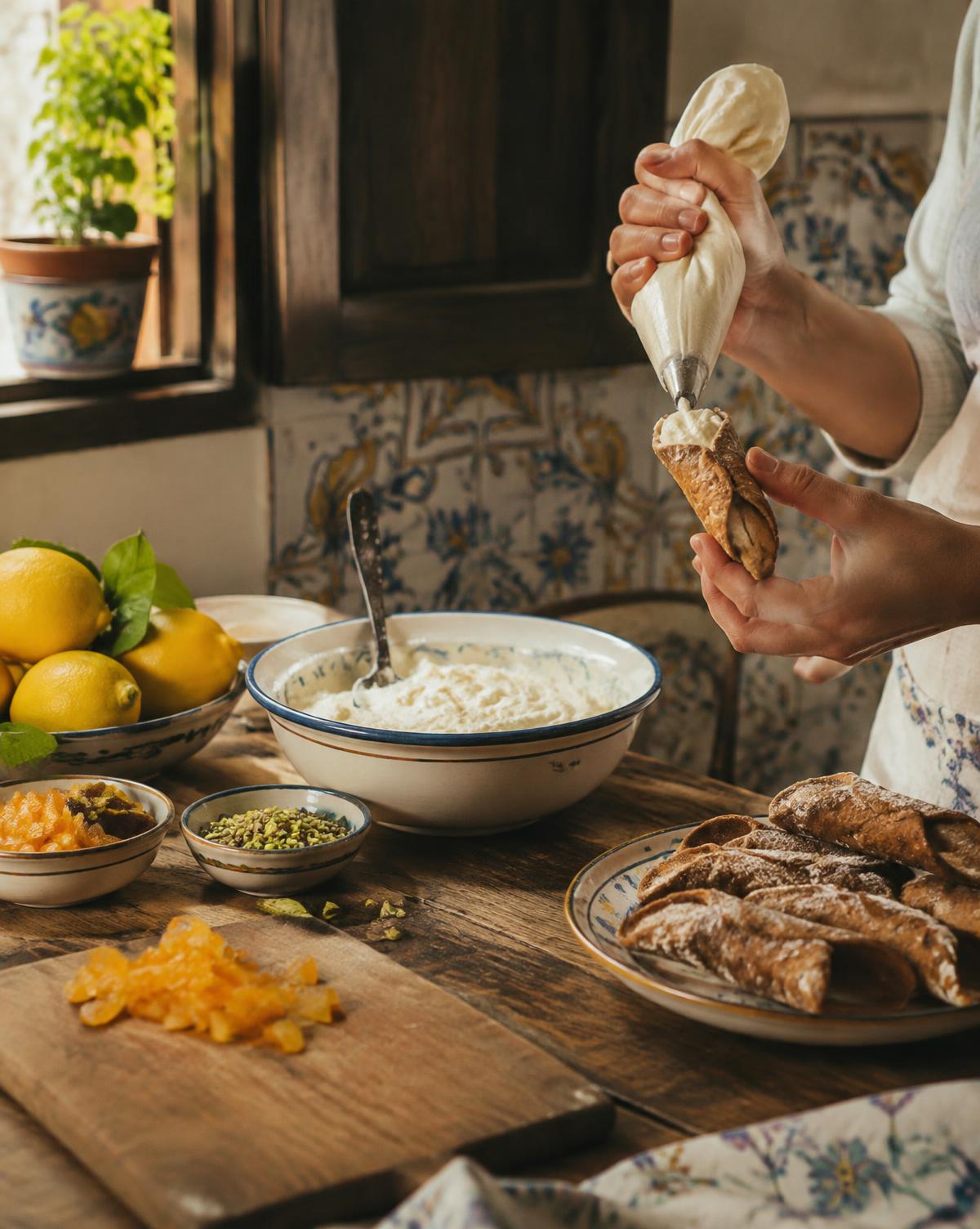 Hands filling cannoli with ricotta beside lemons and pistachios in a tiled kitchen