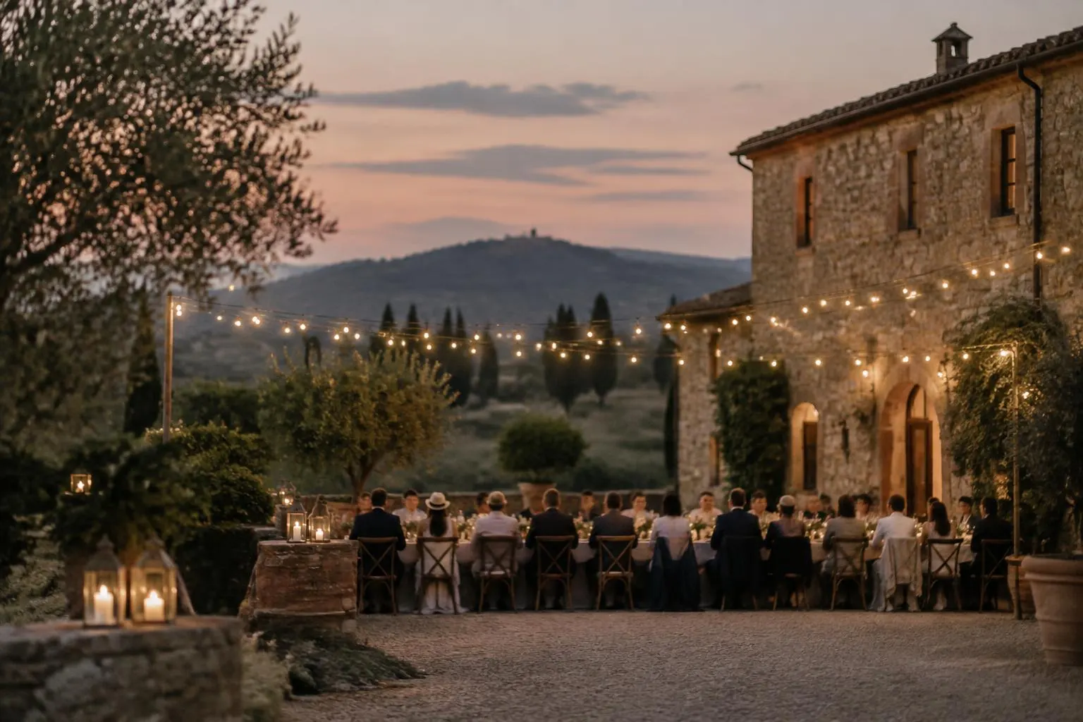 Guests seen from behind at a long dinner table under string lights in a stone farmhouse courtyard at dusk, with hills beyond